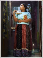 Person wearing Boriby light blue oversized T‑shirt with hand‑painted red sari artwork, paired with Ajrakh patterned skirt, standing indoors near textiles