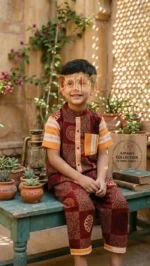 Child in maroon and orange Ajrakh cotton outfit sitting on rustic bench with ethnic craft décor.