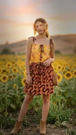 Woman in sleeveless yellow floral top and brown patterned skirt standing in sunflower field at sunset.