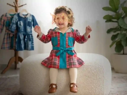 Young girl in a red and blue plaid Boriby dress with turquoise belt and pockets, cream tights, brown shoes, and floral headband, sitting on an ottoman with matching dresses in the background.